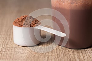 Whey Protein. White scoop with chocolate flavour powder and shaker. Wooden table. Close up, texture