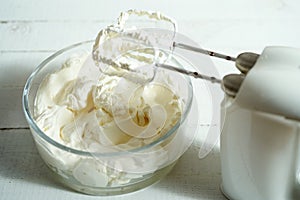 Whipped cream in a bowl and electric hand mixer on bright table.