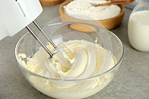 Whipping white cream in glass bowl with mixer on light grey table, closeup