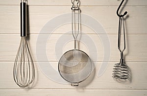 Whisk, masher and strainer hanging against a rustic white wall in restaurant or home kitchen. Kitchen utensils made of