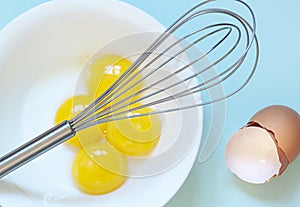 The whisk over fresh raw chicken yolks, eggs shells on the blue background. Baking or food preparation concept
