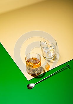 Whiskey glass with bar spoon and empty glass with ice cubes on gre golden background