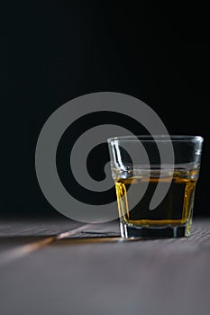 A whiskey glass on the right on a wooden table with a dark background behind. Table in front of a glass in defoc