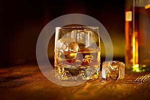 Whiskey with ice cubes. Glass of Whisky and the bottle on wooden table over dark background. Glass of rum alcohol close-up