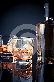 Whiskey with ice in glasses near bottle on black background and light tint blue