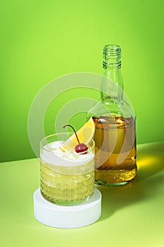 Whiskey sour cocktail with bourbon, lemon juice, egg white and ice, rocks glass and bottle on green background