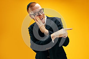 Whispering. Excited young redhead Jewish man in glasses and suit holding hand to mouth against yellow studio background