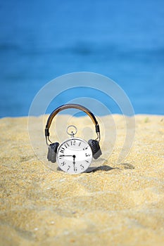 White alarm clock with brown headphones placed on the sand. Beautiful blue sea and sky as a background. Concept for relaxation, tr