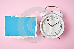 White alarm clock and empty torn paper sheet on a pink background, top view. Time concept