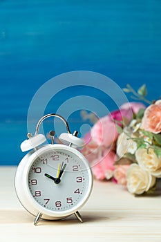 White alarm clock and flowers pink and red rose on wooden table and blue background with copy space for add text and content.