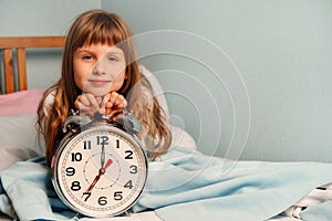 White alarm clock in the hands of a happy smile child girl in the bedroom. 7 o& x27;clock in the morning. Copy space