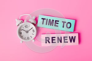 White alarm clock isolated on vivid pink background with text TIME TO RENEW, written on paper notes. Closeup of a personal agenda