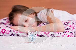 White alarm clock stands on the table on the background of a sleeping girl