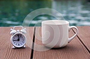 White alarm clock on wooden floor with coffee cup. Clock set at 9 o ' clock.