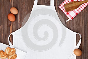 Blank white apron template on wooden table with cookies and eggs, copy space. Kitchen, cooking clothing mockup photo