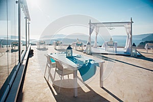 White arch for a wedding ceremony in the open air with lanterns and a platform with white furniture on the background of the sea,