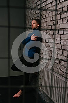 white attractive man in a blue sweater and black pants, barefoot stands near a stone wall, behind bars, a prisoner