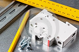 White automatic circuit breaker lying on shiny metal mounting plate of electrical panel with yellow pencil and ruler. Electrical