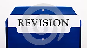 On a white background, a blue folder for papers. In the folder is a sheet of paper with the word REVISION