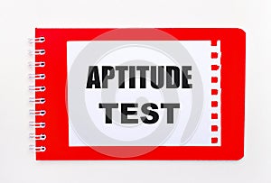 On a white background - a bright red notebook on a spiral. On it is a white sheet of paper with the text APTITUDE TEST