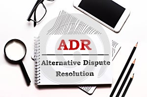 On a white background glasses, a magnifier, pencils, a smartphone and a notebook with the text ADR Alternative Dispute Resolution