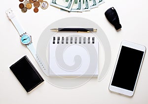 On a white background lie the car key, cash, smartphone, business card holder, blank notebook and pen. Copy space. Business