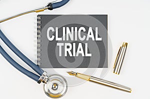 On a white background lie a stethoscope, a pen and a notebook with the inscription - CLINICAL TRIAL