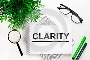 On a white background lies a notebook with the word CLARITY, glasses, a magnifying glass, green markers and a green plant photo