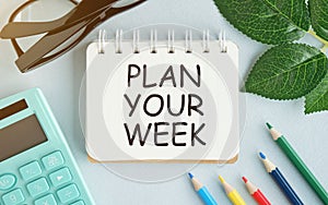 white background lies a notebook with the word PLAN YOUR WEEK, glasses, a magnifying glass, green markers and a green plant