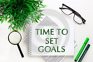 On a white background lies a notebook with words TIME TO SET GOALS, glasses, a magnifying glass, green markers and a green plant