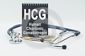 On a white background lies a stethoscope, a pen and a black notebook with the inscription - HCG