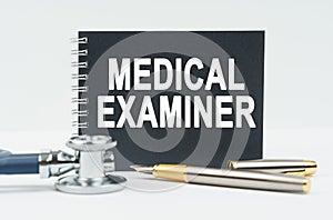 On a white background lies a stethoscope, a pen and a black notebook with the inscription - MEDICAL EXAMINER