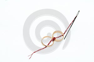 On a white background two gold wedding rings are connected by a red thread coming from a sewing needle