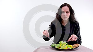 On white background woman in black clothes eats a salad and meat with a fork looks into the frame space for text proper