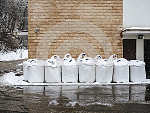 White bags of salt stand in the snow near a wet road against a wall with decorative brickwork.