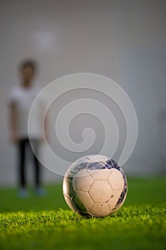 White ball is on the green grass on the football field and blurred litlle boy behind it