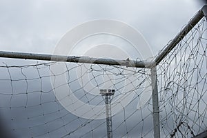White bar of football goal with broken and messy white net seeing a pylon of stadium lighting ready for the night time.