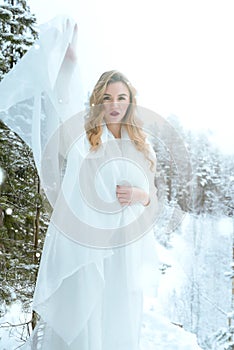 White beautiful young blonde woman in a white dress in winter in the snow in the forest among the trees