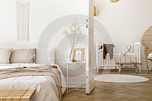 White bedroom interior with bedside table with plant, poster and mug and open door to baby room with rug and white c