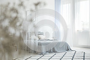 White bedroom interior with carpet, window with drapes and king-size bed with canopy in the photo with blurred foreground