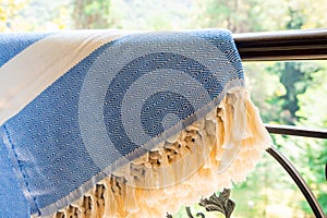 A white beige and blue Turkish peshtemal / towel on a wrought iron railings with blurry nature in the background.