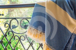 A white beige and blue Turkish peshtemal / towel on a wrought iron railings with blurry nature in the background.