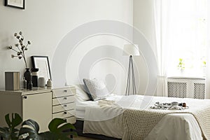 A white and beige color bedroom interior with a bed, a drawer cabinet and a lamp. Pillows, blanket and bedsheets on the bed. Real
