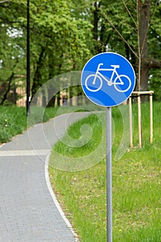 White bicycle lane road traffic sign on blue round background. Bike sign on metal pole, blur narrow street in city park.