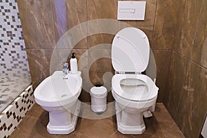 A white bidet, a toilet and a trash can in the bathroom.