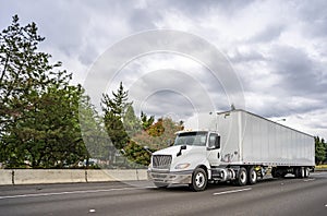 White big rig day cab semi truck with roof spoiler transporting goods in dry van semi trailer driving on the multiline highway