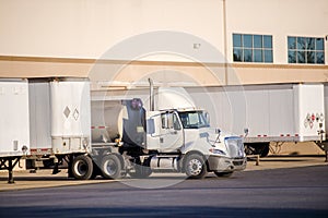 White big rig day cab semi truck with tank semi trailer standing in warehouse dock with another loading semi trailers