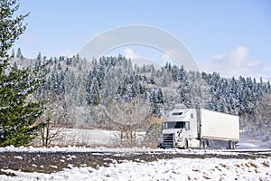 White big rig long haul semi truck with refrigerator semi trailer running on the winding winter snowy road with forest