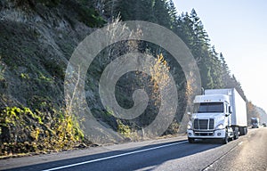 White big rig semi truck with dry van semi trailer driving in front of another semi trucks and cars traffic on wide highway