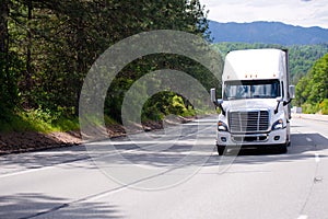 White big rig semi truck with trailer driving by wide multi-line highway with green trees